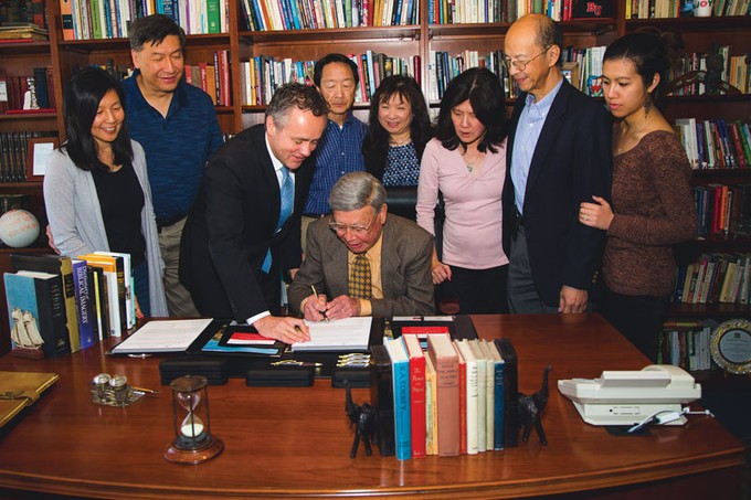 Alton Lim signing a document surrounded by family with Barry Corey standing next to him