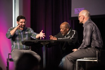 Two men listening to another man talking on a stage